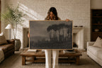 A woman stands in a cozy living room holding up a large framed landscape art print. The artwork depicts a dark, moody forest of tall pine trees under a gray sky.