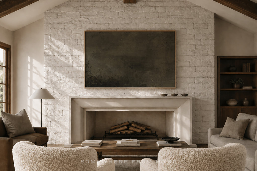 A large dark charcoal wall art canvas displayed in a frame above a white brick fireplace in a rustic, sunlit living room with textured cream boucle armchairs and a wooden coffee table.