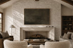 A large dark charcoal wall art canvas displayed in a frame above a white brick fireplace in a rustic, sunlit living room with textured cream boucle armchairs and a wooden coffee table.
