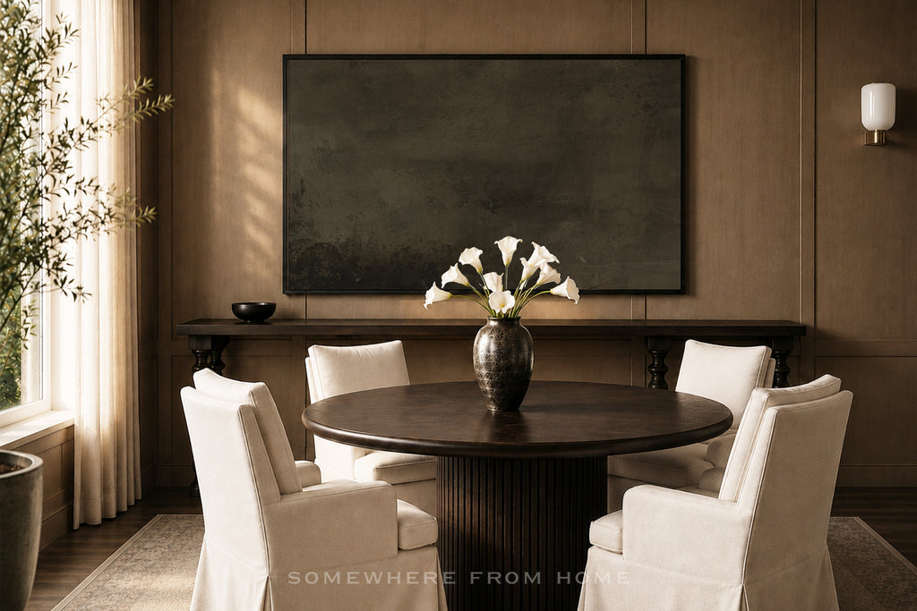 A styled interior scene featuring a round dark wood dining table surrounded by six white upholstered chairs, with a dark vase of white flowers on top. A large dark rectangular wall art piece hangs on a brown paneled wall above a console table.