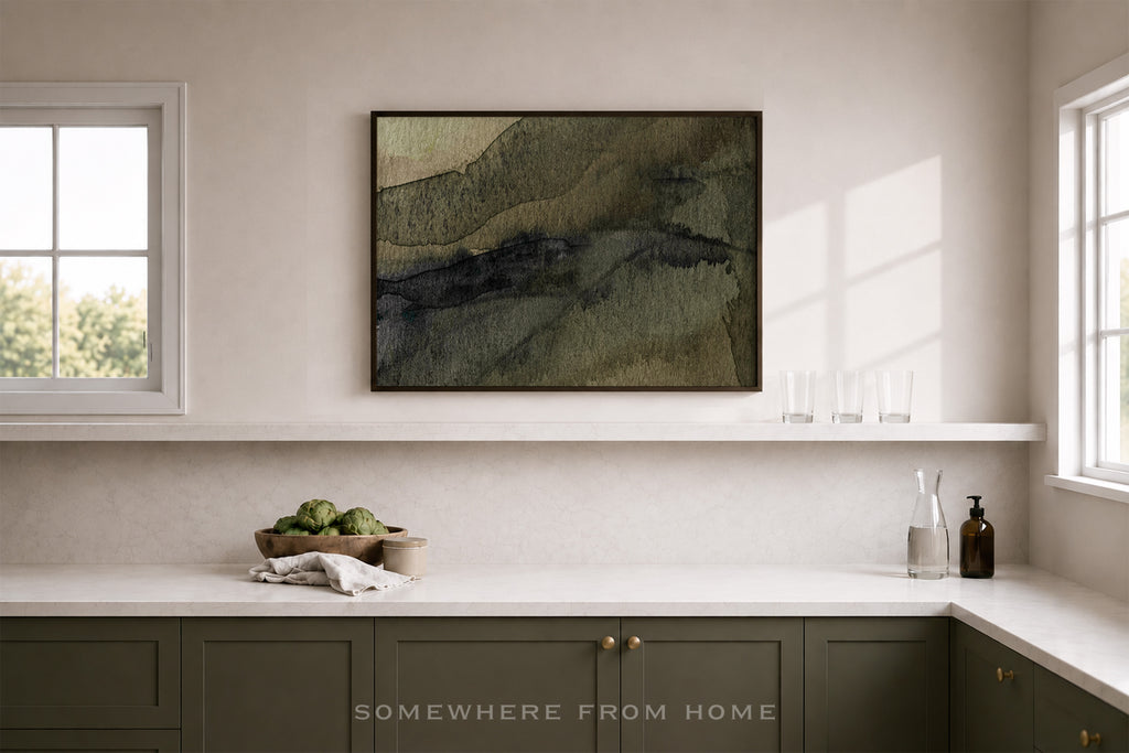 A framed abstract landscape art print with textured olive green, brown, and black brushstrokes, displayed on a white wall above a kitchen counter in a bright, modern kitchen setting.
