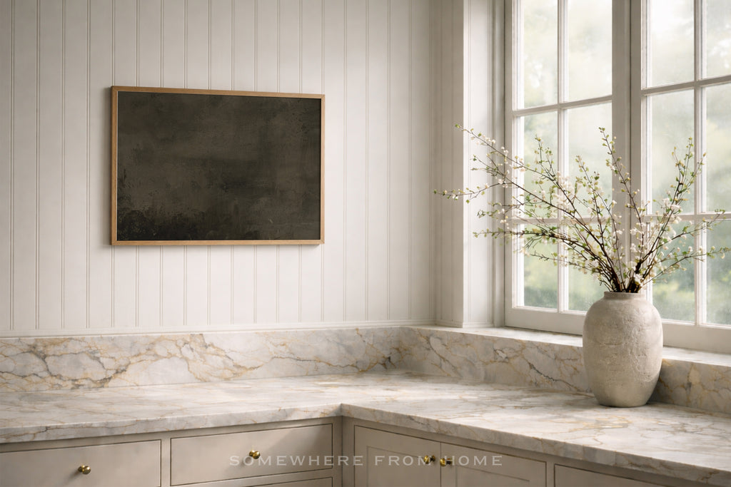 A styled interior corner with white paneled walls, marble countertops, a framed dark artwork on the wall, and a large textured vase with branches on the windowsill.