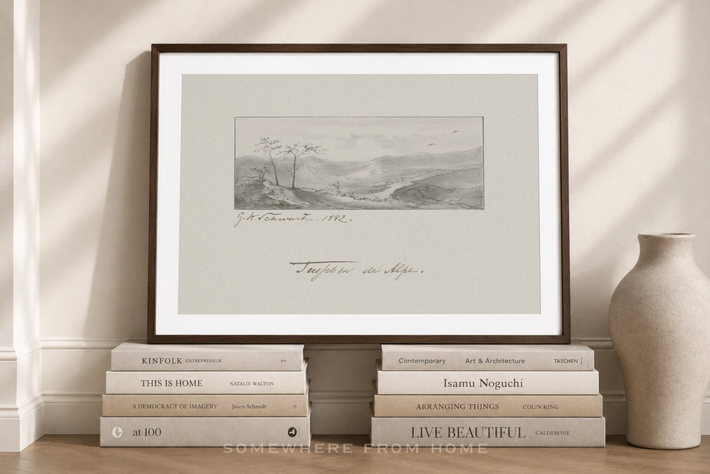 A framed art print featuring a monochrome landscape sketch with cursive writing, styled on a wooden floor against a cream wall with books and a vase.