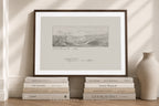 A framed art print featuring a monochrome landscape sketch with cursive writing, styled on a wooden floor against a cream wall with books and a vase.