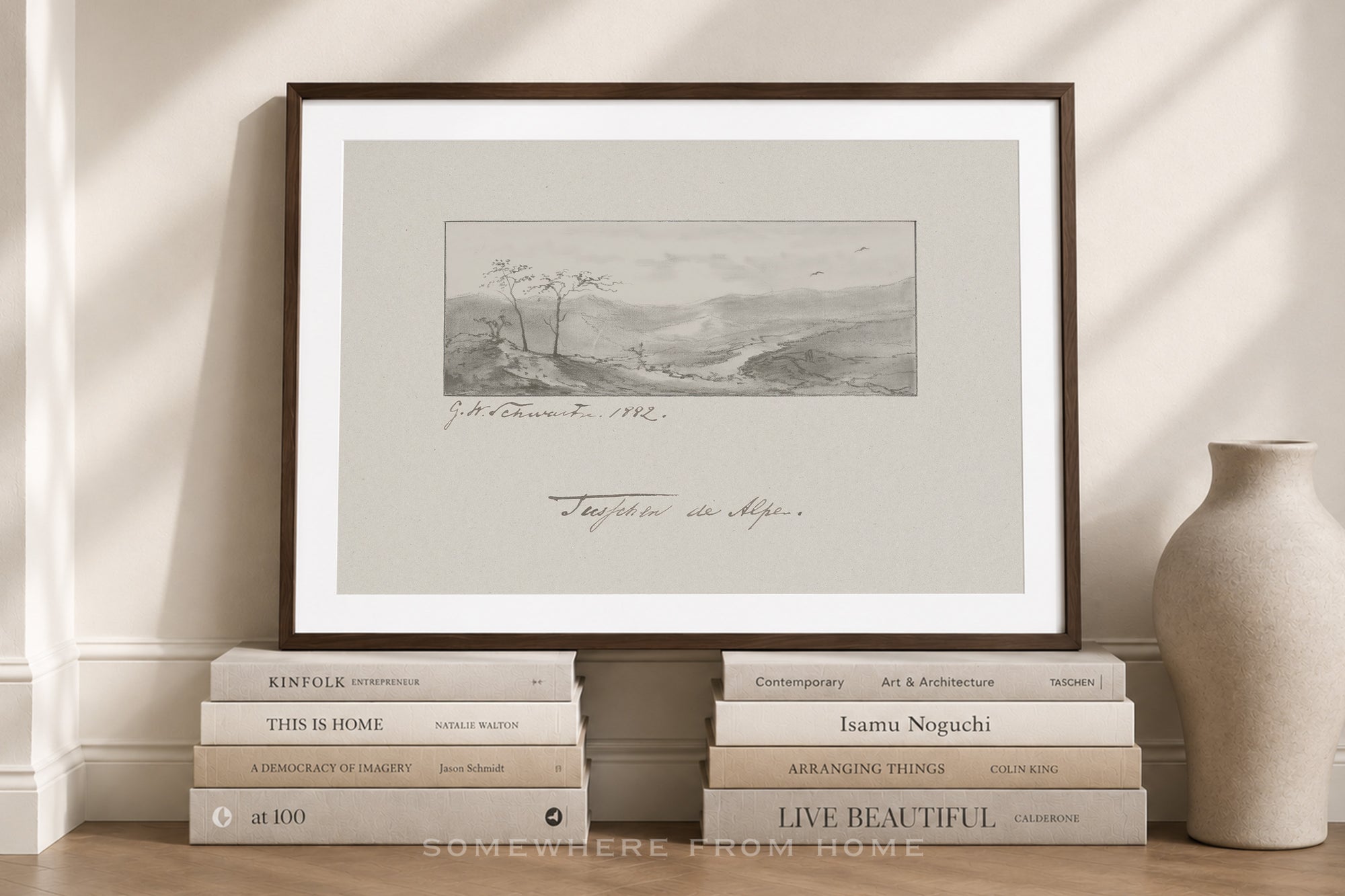 A framed art print featuring a monochrome landscape sketch with cursive writing, styled on a wooden floor against a cream wall with books and a vase.