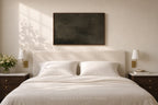 A minimalist bedroom scene featuring a neatly made bed with white linens and pillows, flanked by matching dark wood nightstands with wall-mounted lamps. A large, dark rectangular canvas art piece hangs on the beige wall above the headboard.