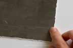 A close-up of a hand holding the edge of a dark gray textured sheet of paper or material against a white background. The sheet has a rough, rustic torn edge and a matte finish.