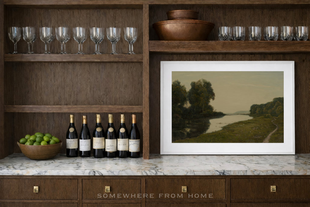 Wine bottles on a marble countertop with a framed landscape painting, surrounded by wine glasses and a bowl of limes.