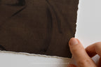 A close-up of a dark brown textured fabric with irregular white tattered edges, held by a person's fingers against a white background. A faint watermark at the bottom reads 'SOMEWHERE FROM HOME'.