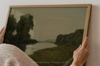 Person holding a framed landscape painting of a river and trees.