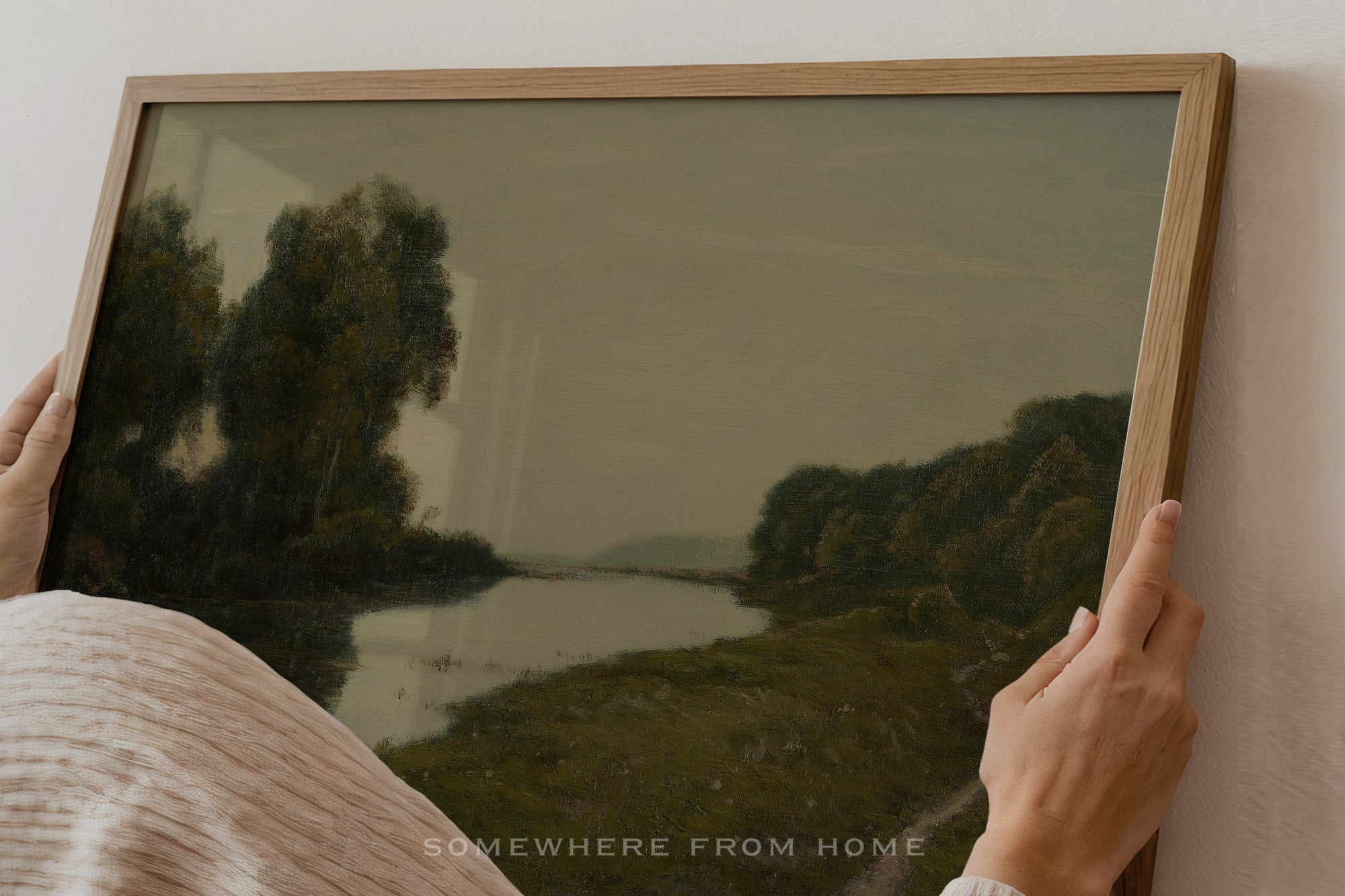 Person holding a framed landscape painting of a river and trees.