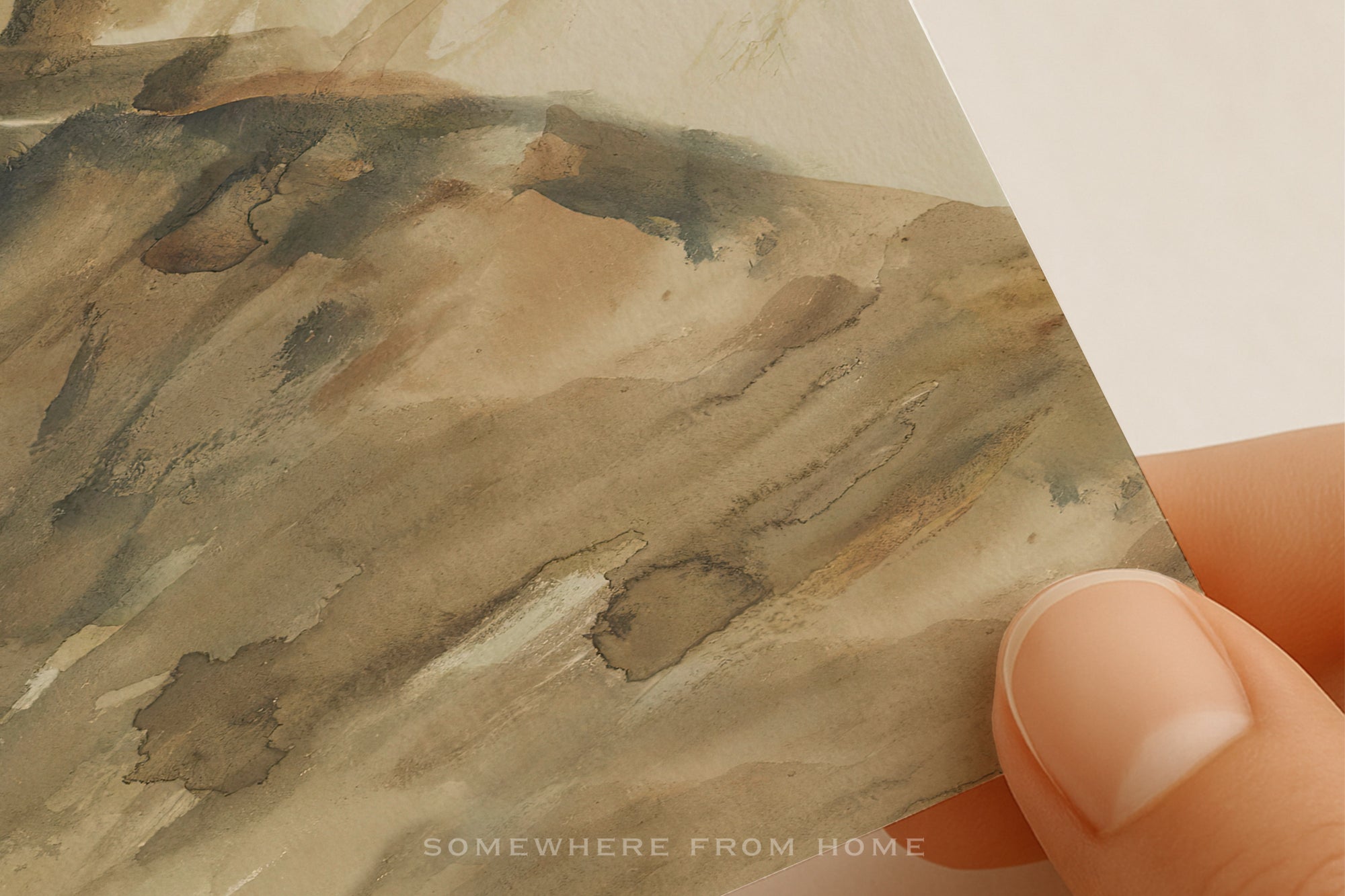 A close-up of a watercolor painting or print showing abstract earthy tones of brown, beige, and gray, with a finger holding the corner of the paper against a white background.