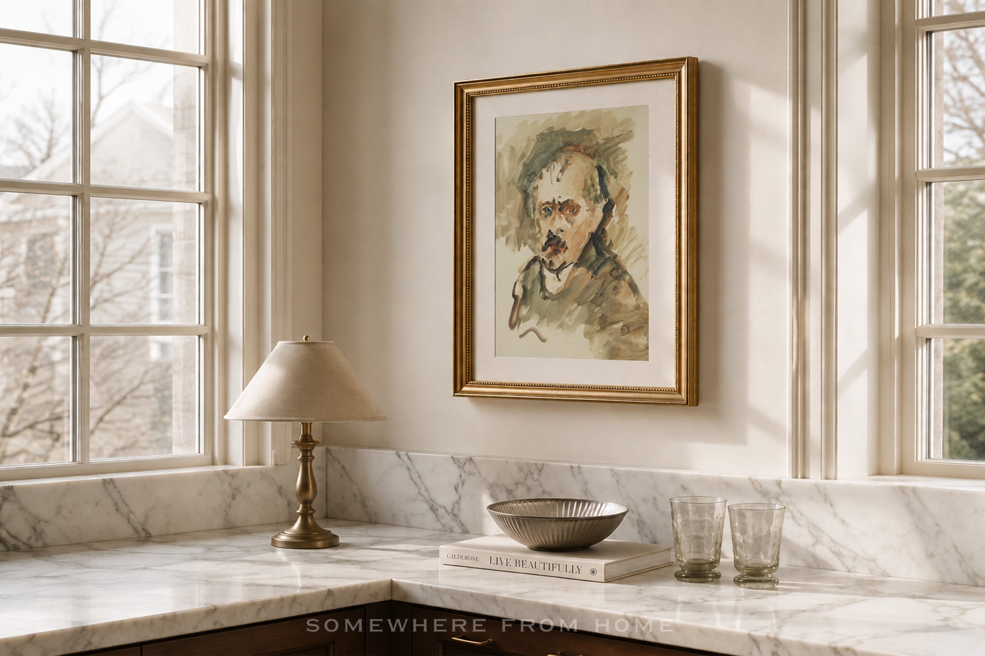 A framed portrait print of a man hangs on a cream wall above a marble countertop in a sunlit room. The artwork has a gold frame and is shown in a styled interior with a lamp, bowl, glasses, and windows.