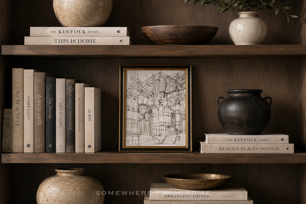 A framed black-and-white architectural sketch print with a gold border displayed on a dark wooden bookshelf, surrounded by books, ceramic vases, and bowls.