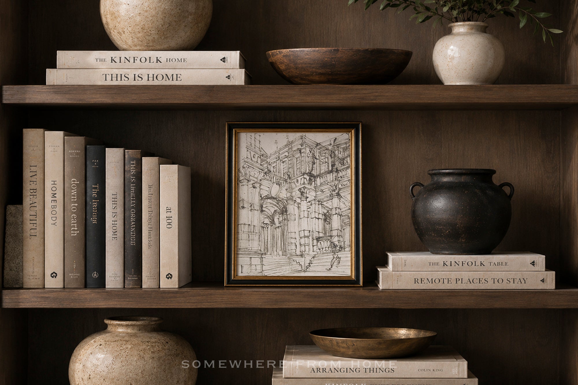 A framed black-and-white architectural sketch print with a gold border displayed on a dark wooden bookshelf, surrounded by books, ceramic vases, and bowls.