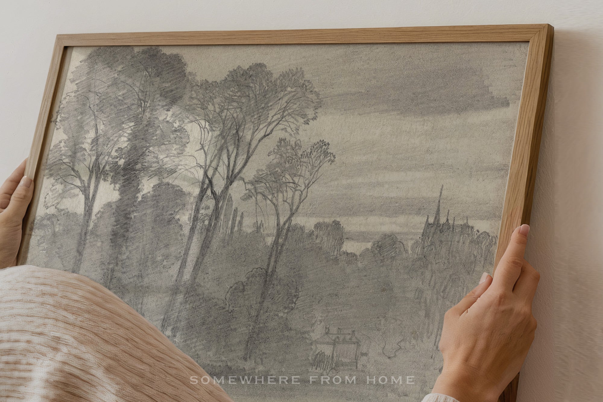 A framed monochrome landscape artwork being held by two hands against a light wall. The print shows trees, a field, and distant buildings in soft gray and black tones, enclosed in a natural wood frame.