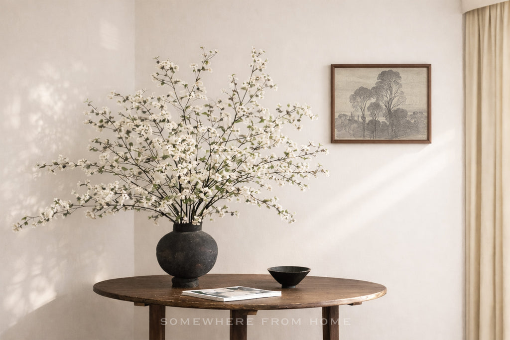A large black ceramic vase filled with white blossoms sits on a round wooden table in a softly lit room. A small black bowl and a magazine rest beside the vase, and a framed landscape print hangs on the wall above.