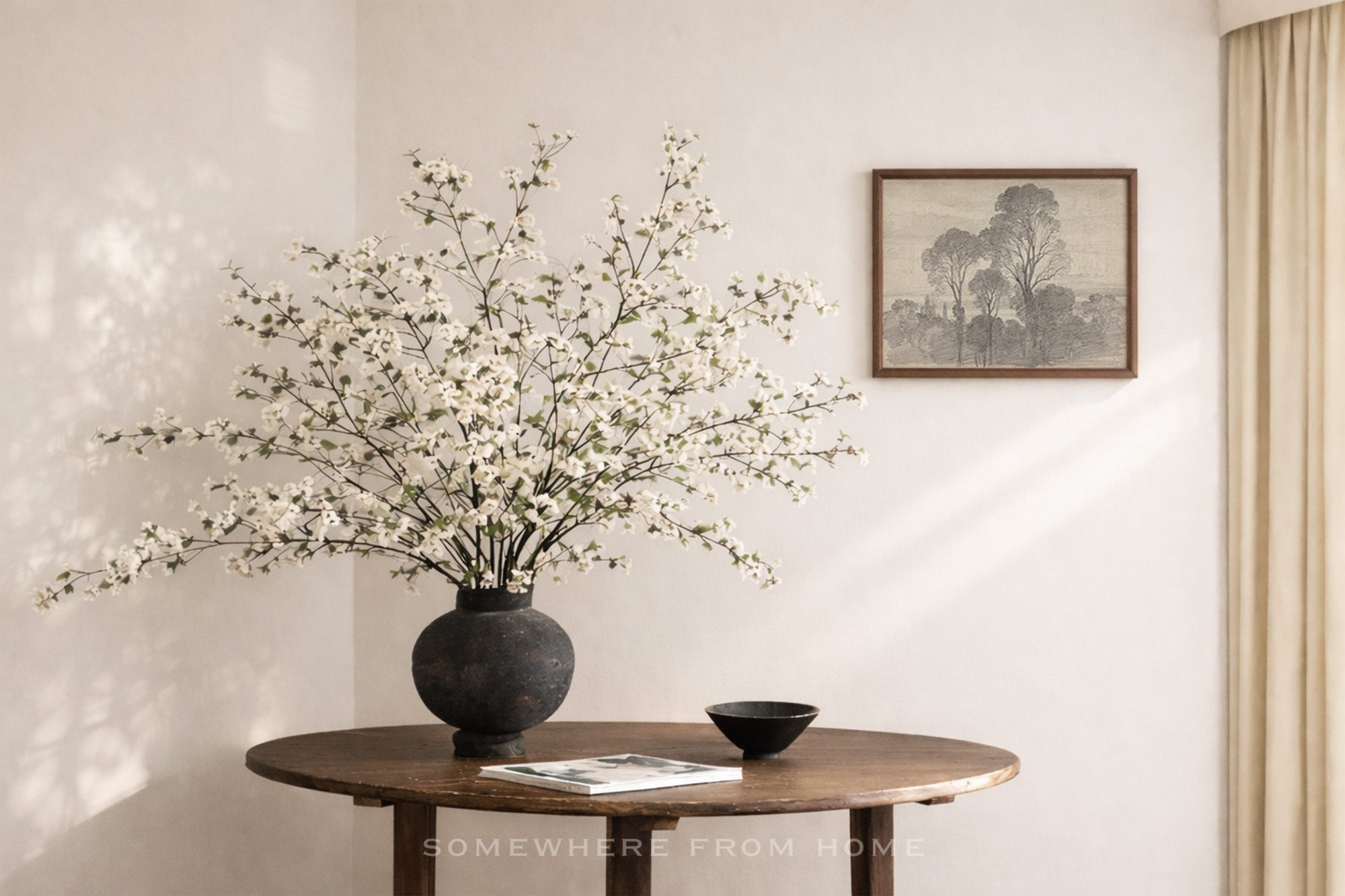 A large black ceramic vase filled with white blossoms sits on a round wooden table in a softly lit room. A small black bowl and a magazine rest beside the vase, and a framed landscape print hangs on the wall above.