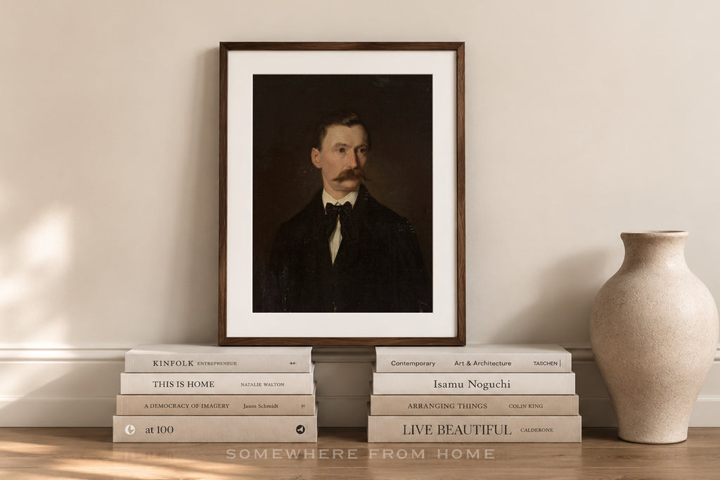 A framed art print featuring a dark portrait of a man with a mustache, styled on a wooden ledge with books and a ceramic vase against a beige wall.