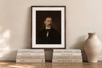 A framed art print featuring a dark portrait of a man with a mustache, styled on a wooden ledge with books and a ceramic vase against a beige wall.