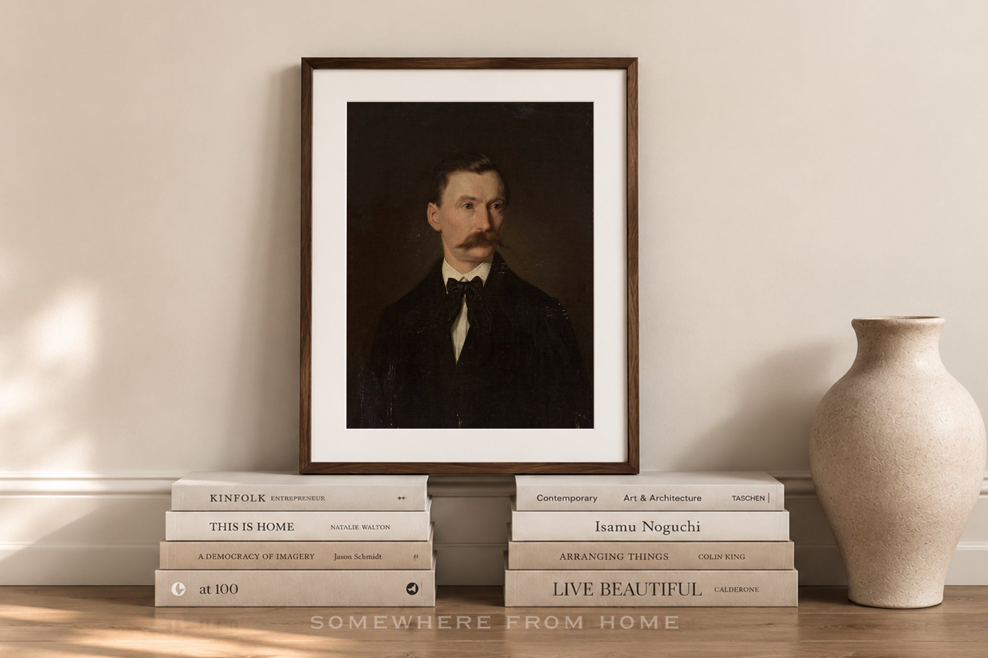 A framed art print featuring a dark portrait of a man with a mustache, styled on a wooden ledge with books and a ceramic vase against a beige wall.