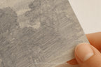 A close-up of a hand holding a square piece of textured gray paper or cardstock with a sketch-like gray wash pattern, against a plain light background.