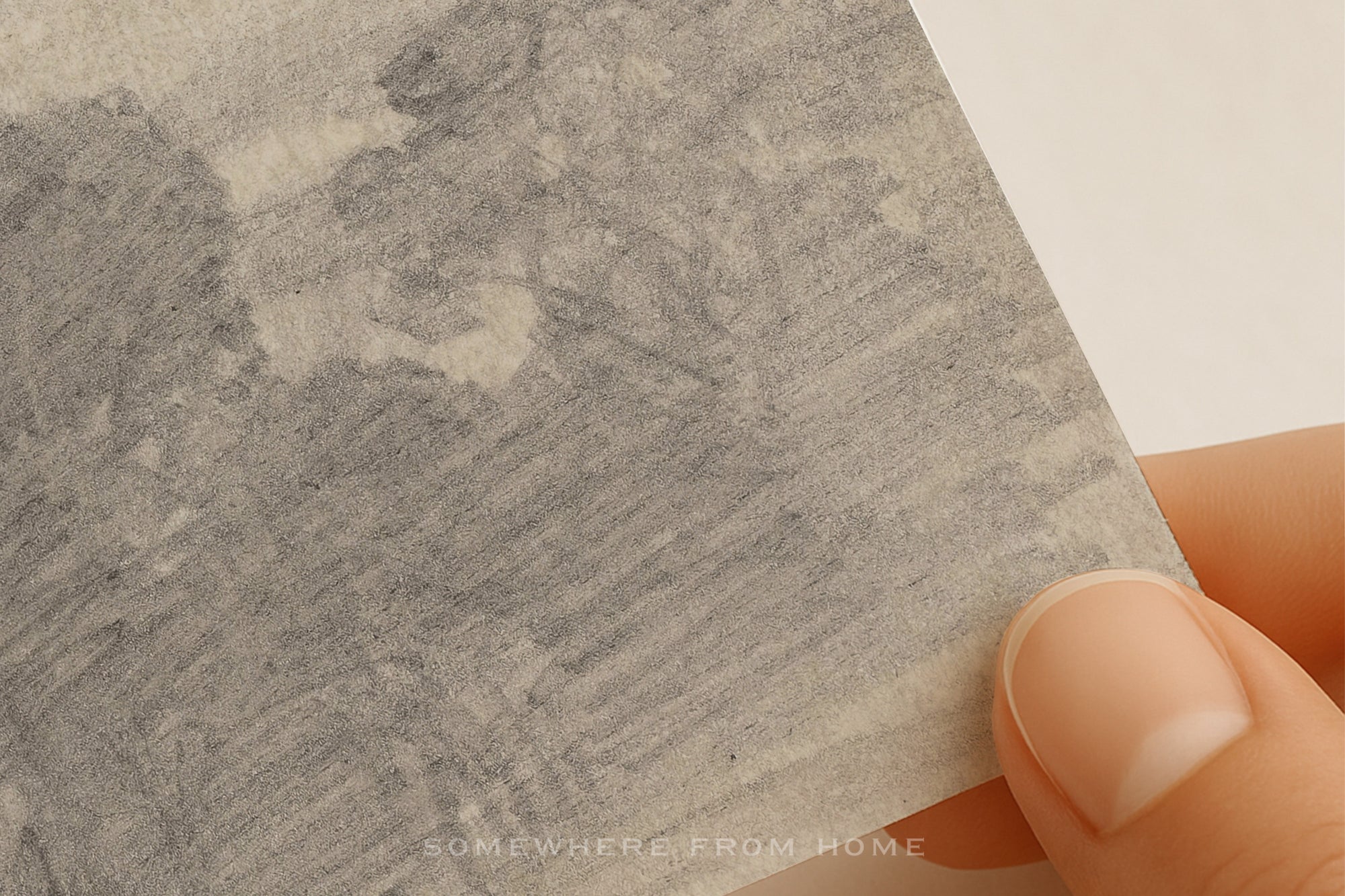 A close-up of a hand holding a square piece of textured gray paper or cardstock with a sketch-like gray wash pattern, against a plain light background.