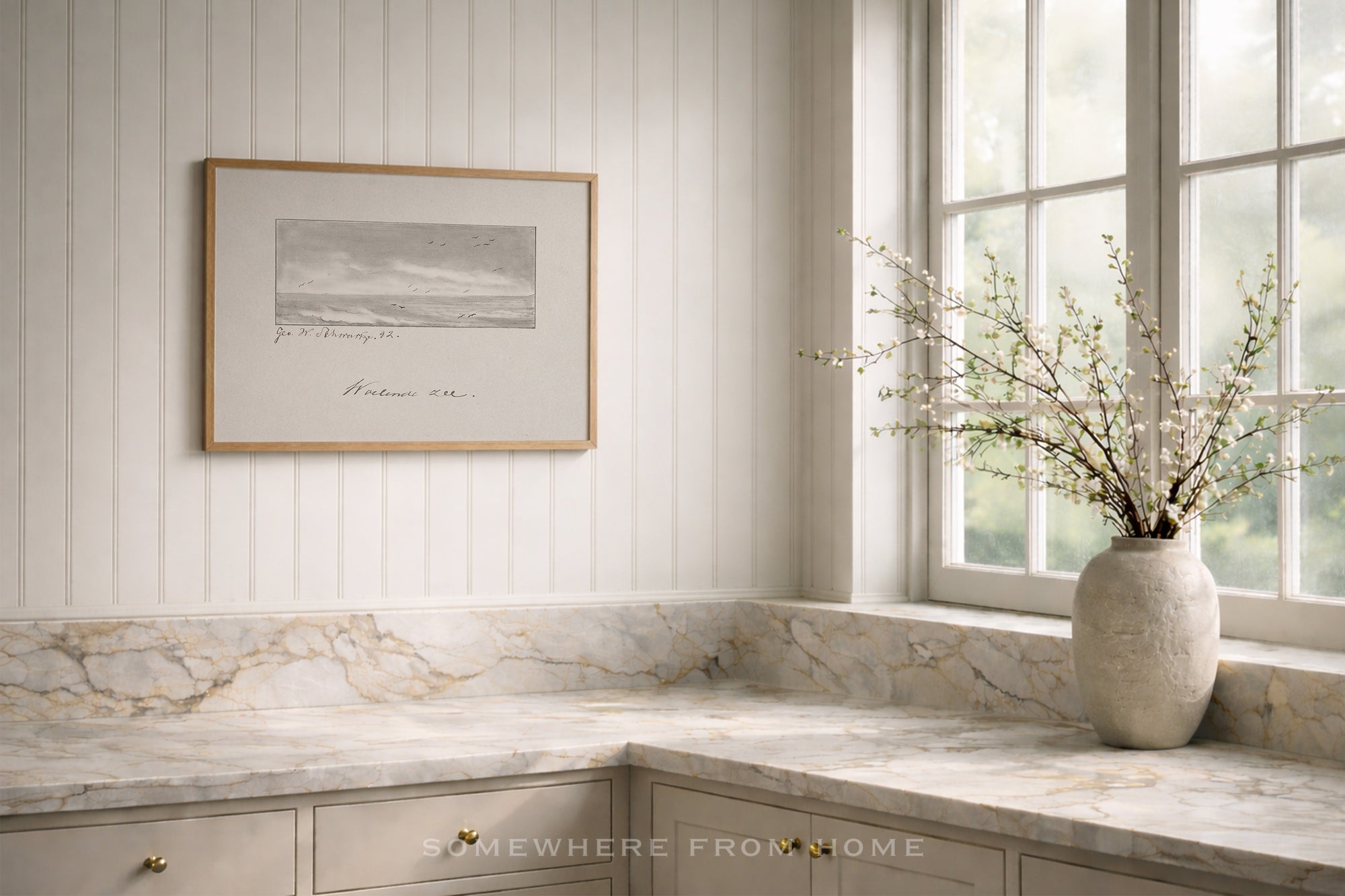 A framed black and white landscape print hangs on a white paneled wall above a marble countertop in a bright kitchen. A textured ceramic vase with delicate branches sits by the window.