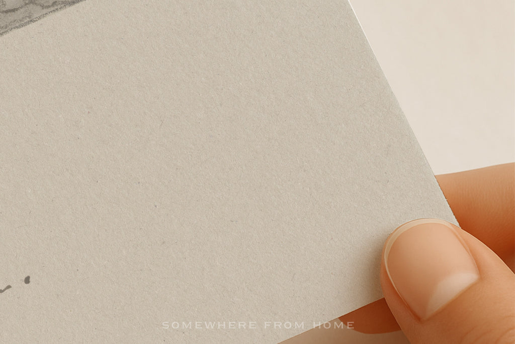 A close-up of a hand holding the corner of a textured off-white paper card or notebook page against a clean white background.