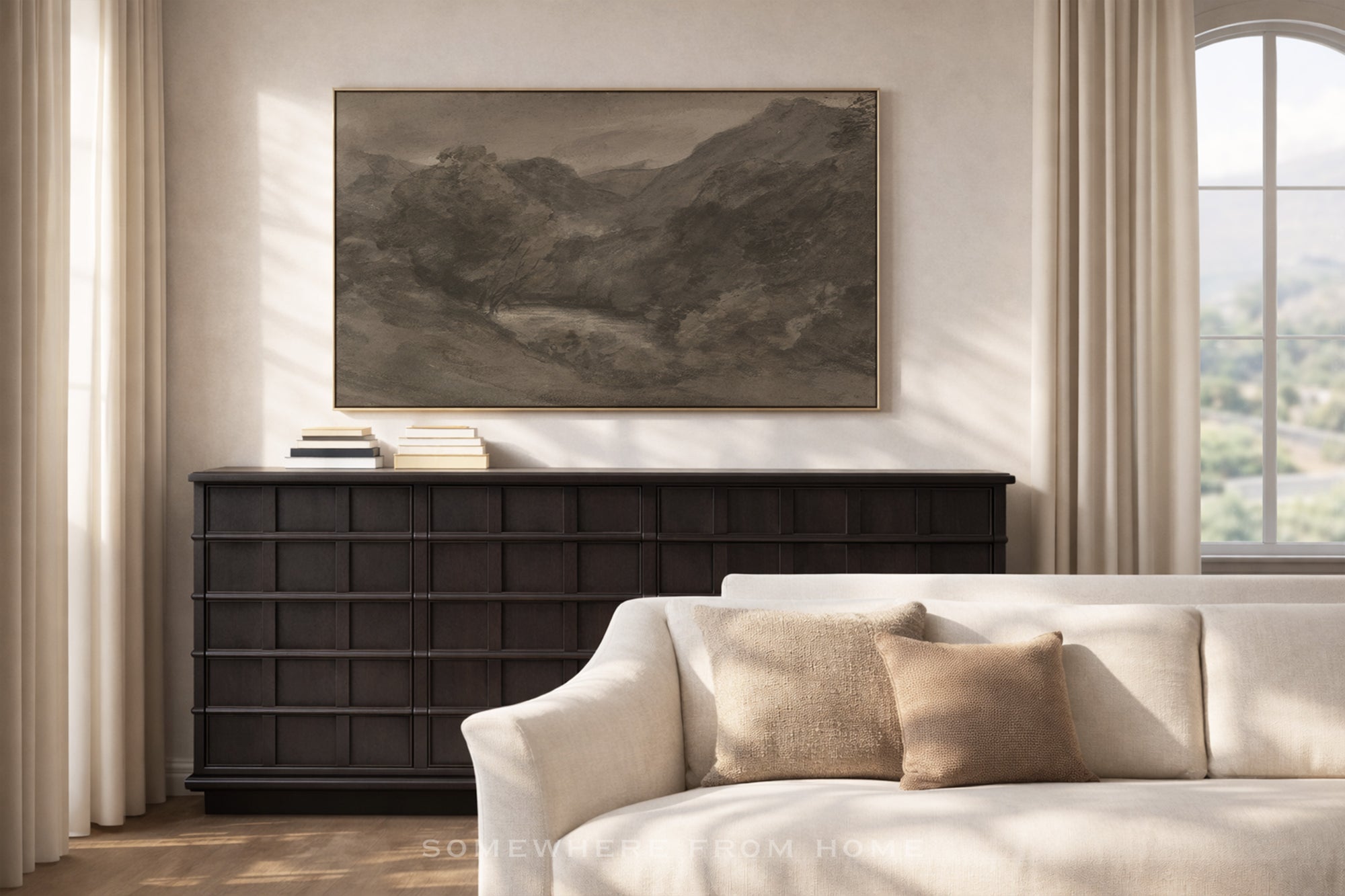 A framed landscape art print in muted earth tones hangs on a beige wall above a dark wood console table in a styled living room with a cream sofa and pillows.