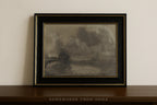 A framed landscape art print featuring a smoky, atmospheric scene in muted gray and brown tones, housed in a black frame with gold trim, resting on a dark wooden shelf against a light beige wall.