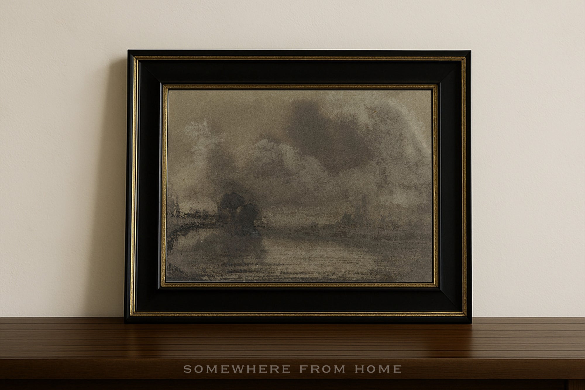 A framed landscape art print featuring a smoky, atmospheric scene in muted gray and brown tones, housed in a black frame with gold trim, resting on a dark wooden shelf against a light beige wall.