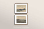 Two framed landscape art prints with white mats and thin brown frames, displayed vertically on a light beige wall. The prints feature soft, muted watercolor-style scenes in gray, blue, and beige tones.