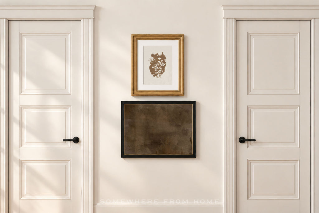 A styled interior scene showing a cream-colored wall between two white paneled doors, with two framed art pieces hanging in the center. The top frame contains a gold-toned print in a gold frame, and the bottom frame contains a dark textured print in a black frame.