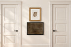A styled interior scene showing a cream-colored wall between two white paneled doors, with two framed art pieces hanging in the center. The top frame contains a gold-toned print in a gold frame, and the bottom frame contains a dark textured print in a black frame.