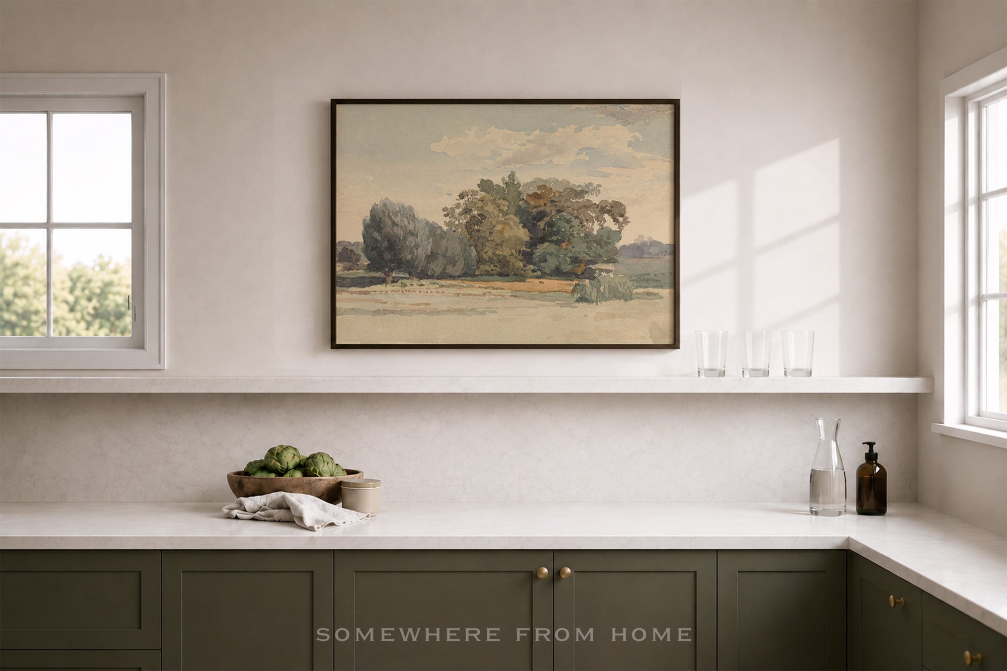 A framed landscape art print hangs on a beige wall above a kitchen counter. The print depicts a tree-lined field under a cloudy sky.