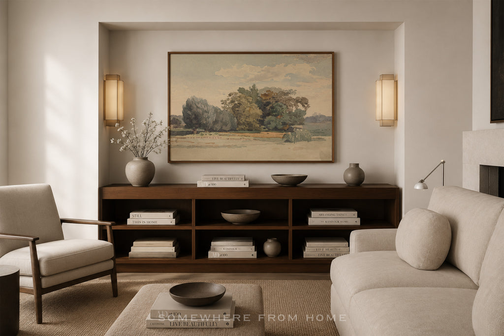 A framed landscape painting hangs on a beige wall above a dark wood console in a styled living room with a cream sofa, armchair, and decorative vases.