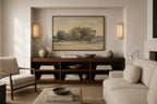 A framed landscape painting hangs on a beige wall above a dark wood console in a styled living room with a cream sofa, armchair, and decorative vases.