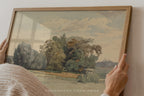 A framed landscape painting being held by two hands against a light-colored wall. The painting depicts trees, a field, and a cloudy sky, enclosed in a light wood frame.