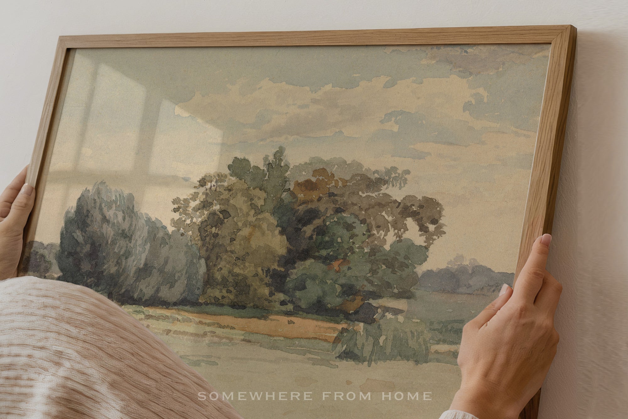 A framed landscape painting being held by two hands against a light-colored wall. The painting depicts trees, a field, and a cloudy sky, enclosed in a light wood frame.
