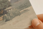 A close-up of a hand holding a square art print or card showing a watercolor landscape with muted gray, green, beige, and brown tones. The text 'SOMEWHERE FROM HOME' appears faintly at the bottom.