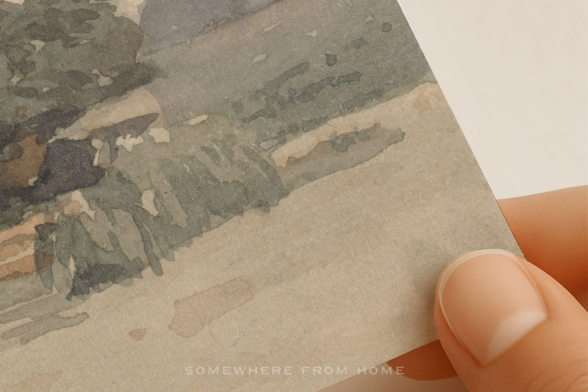 A close-up of a hand holding a square art print or card showing a watercolor landscape with muted gray, green, beige, and brown tones. The text 'SOMEWHERE FROM HOME' appears faintly at the bottom.