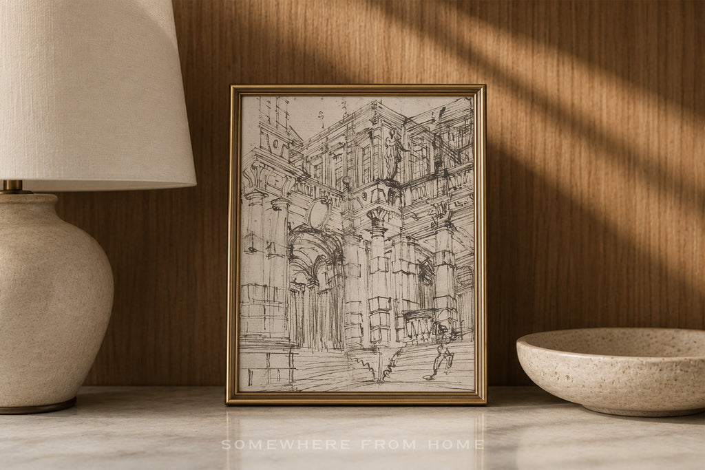 A framed black and white architectural sketch print displayed on a marble surface against a wood-paneled wall, flanked by a lamp on the left and a ceramic bowl on the right.