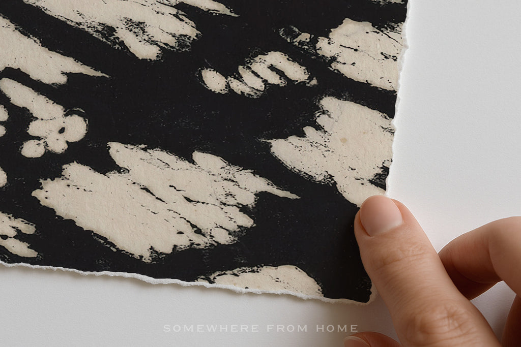A close-up of a hand holding the edge of a black and off-white patterned abstract art print with deckled edges against a plain white background. 