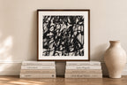 A framed abstract black-and-white art print leans against a beige wall on a wooden floor, styled with books and a ceramic vase. The print features bold, energetic black brushstrokes on a white background, enclosed in a dark brown wood frame.