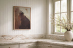 A framed abstract painting with earthy brown, beige, and dark tones hangs on a white paneled wall above a marble countertop in a bright kitchen. A textured vase with small blossoms sits by the window.