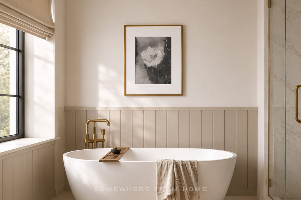 A modern freestanding white bathtub in a softly lit bathroom with beige wainscoting, a gold faucet, a framed black-and-white floral print on the wall, and a glass shower door on the right.