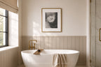 A modern freestanding white bathtub in a softly lit bathroom with beige wainscoting, a gold faucet, a framed black-and-white floral print on the wall, and a glass shower door on the right.