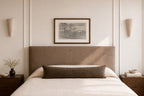 A styled bedroom scene featuring a bed with a tall upholstered headboard, white bedding, a dark brown lumbar pillow, matching bedside tables, wall sconces, and a framed landscape print on the wall.