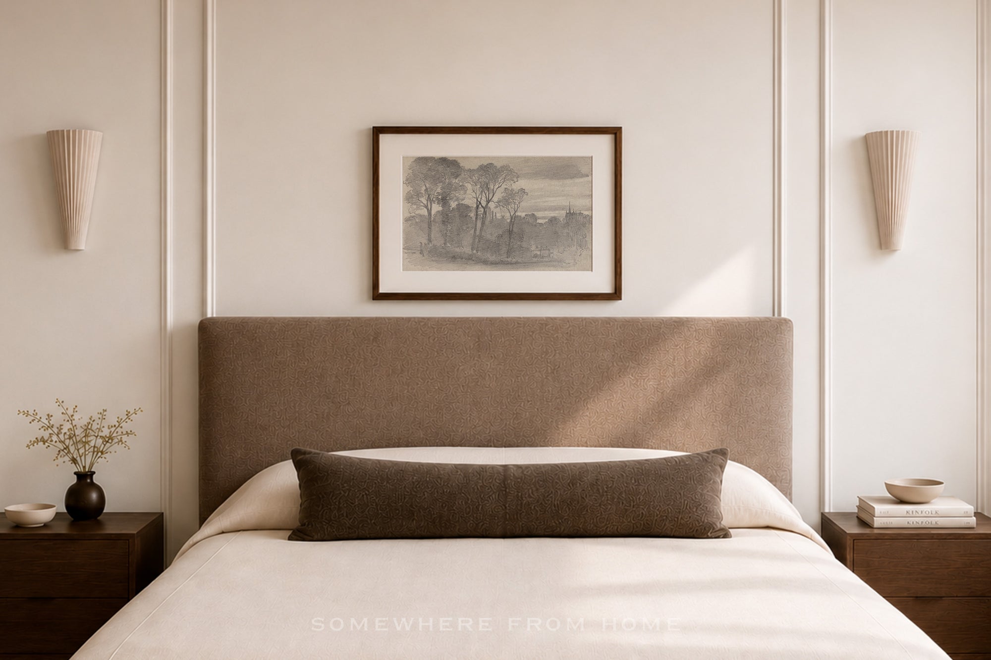 A styled bedroom scene featuring a bed with a tall upholstered headboard, white bedding, a dark brown lumbar pillow, matching bedside tables, wall sconces, and a framed landscape print on the wall.
